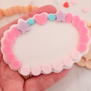 May include: A white, heart-shaped cookie cutter with a bracelet design. The bracelet is decorated with pink and light purple stars, hearts, and beads. Additional cookie cutters and heart-shaped sprinkles are also visible.