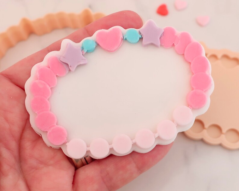 May include: A white, heart-shaped cookie cutter with a bracelet design. The bracelet is decorated with pink and light purple stars, hearts, and beads. Additional cookie cutters and heart-shaped sprinkles are also visible.