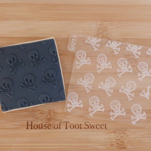 May include: A square biscuit with a grey skull and crossbones pattern next to a clear plastic sheet with the same design. The biscuit is on a wooden surface. The text "House of Toot Sweet" is visible.