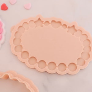 May include: A light peach-colored cookie cutter with a scalloped edge and a central oval shape. The cutter has decorative cutouts of stars and hearts. Several small heart-shaped candies are scattered nearby.
