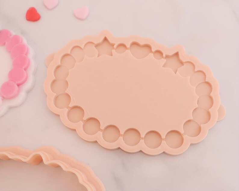 May include: A light peach-colored cookie cutter with a scalloped edge and a central oval shape. The cutter has decorative cutouts of stars and hearts. Several small heart-shaped candies are scattered nearby.
