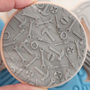 May include: A round biscuit featuring a grey tool pattern, including hammers, spanners, and screwdrivers. The biscuit has a light brown edge and is held by a hand. The tools are embossed on the grey surface.