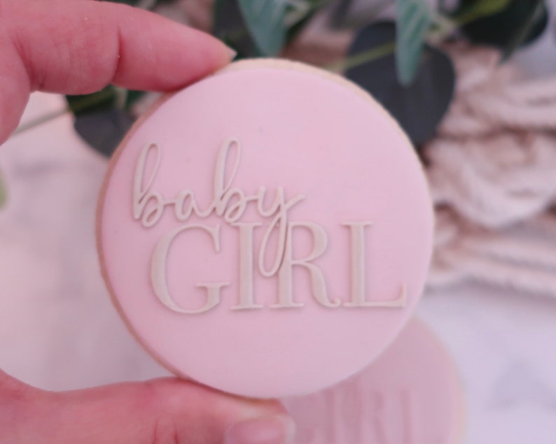 May include: A round, light pink cookie with the words "baby GIRL" in a lighter shade of pink. The cookie is held by a hand, with a blurred background of greenery and a white woven texture.