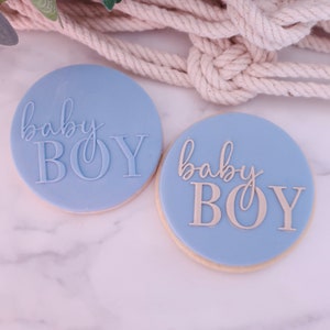 May include: Two round, light blue cookies with the words "baby BOY" in a cursive and block font. The cookies are on a white marble surface, with a beige rope and greenery in the background.