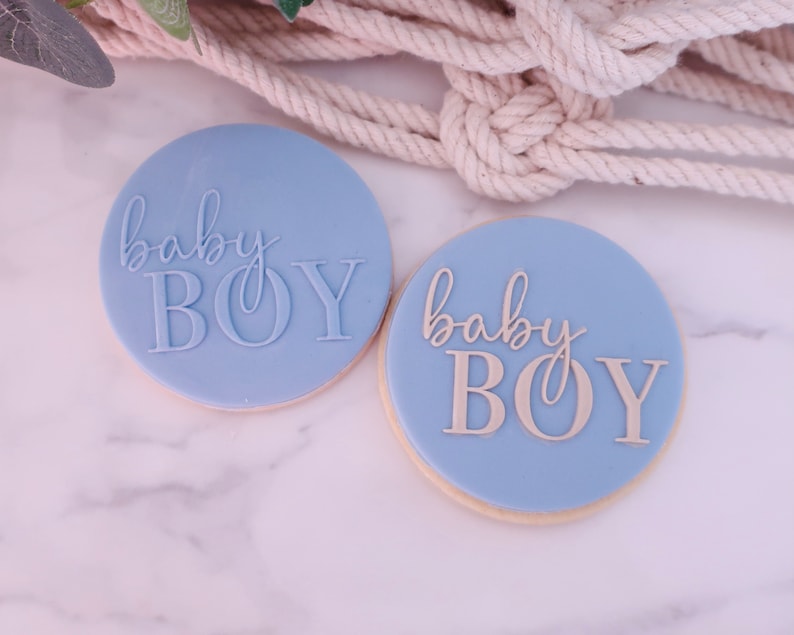 May include: Two round, light blue cookies with the words "baby BOY" in a cursive and block font. The cookies are on a white marble surface, with a beige rope and greenery in the background.