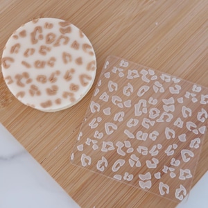 May include: A circular biscuit with brown leopard print spots is placed beside a clear, square stamp featuring a similar leopard print design. Both items rest on a wooden surface.