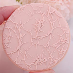 May include: A round, light pink cookie with a detailed floral pattern. The design features embossed flowers and leaves, creating a delicate, textured surface. The cookie has a light brown edge.