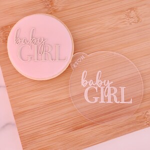 May include: A round, light pink cookie with the words "baby GIRL" embossed in a cursive and block font. A clear acrylic stamp with the same text is next to the cookie. The items are on a wooden surface.