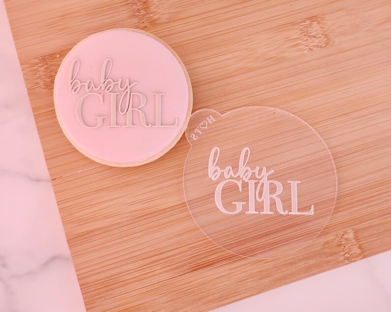 May include: A round, light pink cookie with the words "baby GIRL" embossed in a cursive and block font. A clear acrylic stamp with the same text is next to the cookie. The items are on a wooden surface.