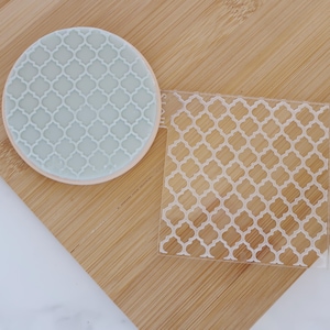 Könnte beinhalten: Ein runder, hellblauer Keks mit einem weißen Vierpassmuster, neben einer transparenten quadratischen Schablone mit dem gleichen Design. Die Artikel liegen auf einem Holzschneidebrett. Der Keks hat einen hellbraunen Rand.