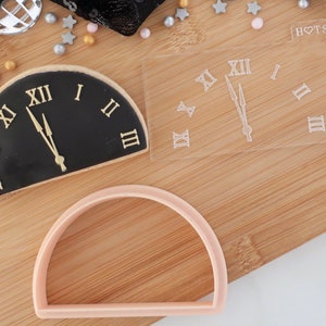 May include: A New Year's Eve themed cookie with a clock face design, featuring Roman numerals and hands pointing to midnight. A clear plastic stencil and a pink cookie cutter are also present, all on a wooden surface.