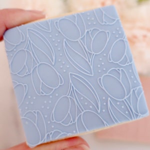 May include: A light blue, square cookie stamp with a floral tulip design. The stamp features a repeating pattern of tulip flowers and leaves, with small dot accents. The design is raised, creating a 3D effect. The stamp is held by a hand.