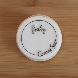 K&ouml;nnte beinhalten: Ein runder Keks mit wei&szlig;er Glasur und den Worten "Baby Coming Soon" in schwarzer Schrift. Der Keks liegt auf einer Holzoberfl&auml;che.