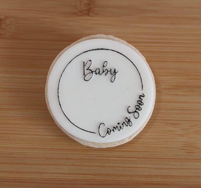 K&ouml;nnte beinhalten: Ein runder Keks mit wei&szlig;er Glasur und den Worten "Baby Coming Soon" in schwarzer Schrift. Der Keks liegt auf einer Holzoberfl&auml;che.