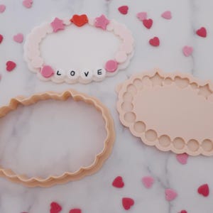 May include: Three pink and peach-colored cookie cutters and a decorative bracelet with the word "LOVE" spelled out. The bracelet has pink stars and a red heart. Scattered around are heart-shaped sprinkles.