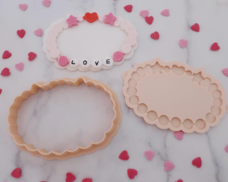 May include: Three pink and peach-colored cookie cutters and a decorative bracelet with the word "LOVE" spelled out. The bracelet has pink stars and a red heart. Scattered around are heart-shaped sprinkles.