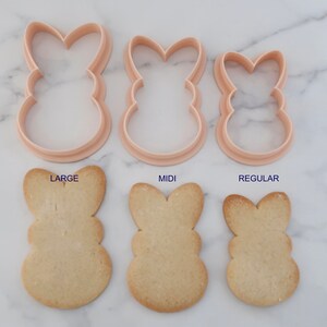 May include: Three bunny-shaped cookie cutters in varying sizes, with corresponding baked cookies below. The cutters are light pink, and the cookies are golden brown. Text labels read "LARGE", "MIDI", and "REGULAR".