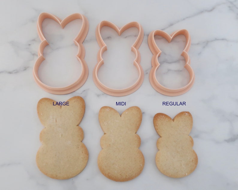 May include: Three bunny-shaped cookie cutters in varying sizes, with corresponding baked cookies below. The cutters are light pink, and the cookies are golden brown. Text labels read "LARGE", "MIDI", and "REGULAR".