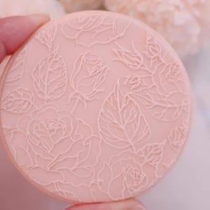 May include: A round, light pink object with a detailed floral design of roses and leaves. The raised pattern is in a lighter shade of pink, creating a subtle contrast. The object is held by a hand, with soft pink flowers in the background.