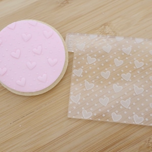 May include: A pink cookie with heart-shaped imprints and a clear plastic stencil featuring a heart and dot design. The cookie is round and placed beside the stencil on a wooden surface.