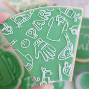 May include: A green cookie decorated with white golf-themed illustrations, such as a golf shirt and golf ball, and the word "PAR". The cookie is held by fingers. Other cookies are visible in the background.
