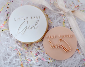 May include: A round cookie with the words "LITTLE BABY Girl" in a light gray color. Next to it is a round stamp with the same text in reverse. The items are on a bed of white paper and colorful confetti.