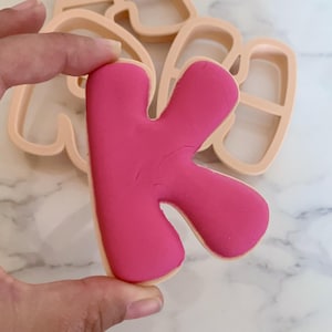 Könnte beinhalten: Ein leuchtend pinker Keks in Form des Buchstabens K mit glatter Glasur, gehalten vor einem hellpfirsichfarbenen Kunststoff-Ausstecher in Form desselben Buchstabens. Der Keks hat ein abgerundetes Bubble-Schrift-Design mit einer soliden, magentafarbenen Oberfläche.
