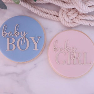 May include: Two round cookies, one light blue and one pink, with the words "baby BOY" and "baby GIRL" in a cursive font. The cookies are on a marble surface, with a rope in the background.