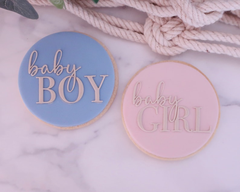 May include: Two round cookies, one light blue and one pink, with the words "baby BOY" and "baby GIRL" in a cursive font. The cookies are on a marble surface, with a rope in the background.