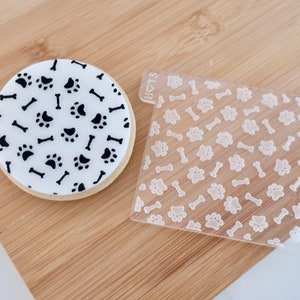 May include: A round cookie decorated with black paw prints and bones on a white background. A clear plastic stencil with the same pattern is next to the cookie. The cookie and stencil are on a wooden surface.
