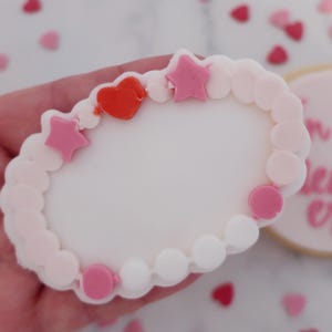 May include: A Valentine's Day-themed cookie shaped like a bracelet, decorated with pink and white icing. The bracelet features pink star and circle accents, and a red heart. Additional heart-shaped sprinkles are scattered around.