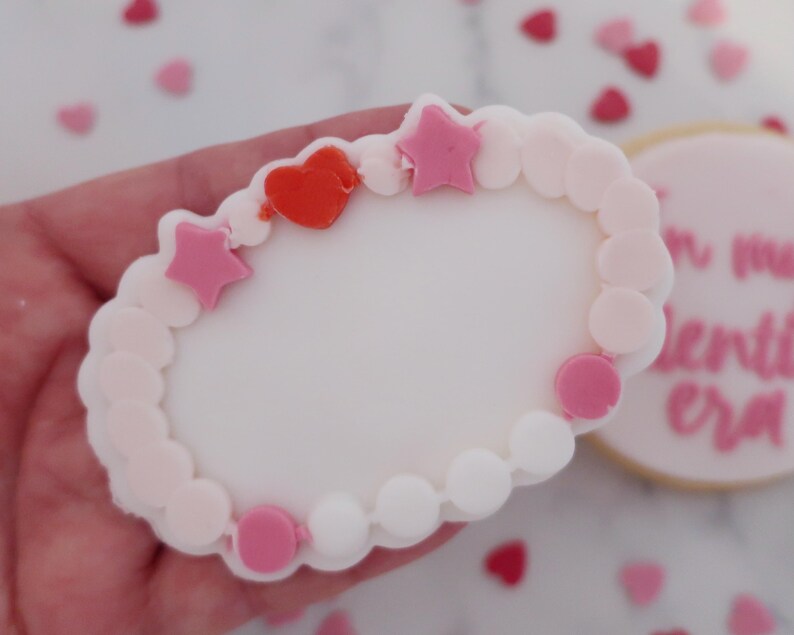 May include: A Valentine's Day-themed cookie shaped like a bracelet, decorated with pink and white icing. The bracelet features pink star and circle accents, and a red heart. Additional heart-shaped sprinkles are scattered around.