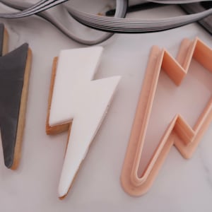May include: Three lightning bolt-shaped items. One is a grey cookie with grey icing, another is a white cookie with white icing, and the third is a pink cookie cutter. Ribbons are in the background.
