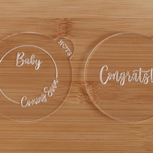 K&ouml;nnte beinhalten: Zwei transparente Acrylscheiben f&uuml;r eine Babyank&uuml;ndigung. Eine Scheibe sagt "Baby Coming Soon" in einem Kreis. Die andere sagt "Congrats!!!". Beide haben eine herzf&ouml;rmige Lasche mit dem Wort "HOTS".