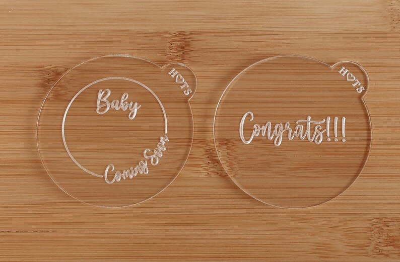 K&ouml;nnte beinhalten: Zwei transparente Acrylscheiben f&uuml;r eine Babyank&uuml;ndigung. Eine Scheibe sagt "Baby Coming Soon" in einem Kreis. Die andere sagt "Congrats!!!". Beide haben eine herzf&ouml;rmige Lasche mit dem Wort "HOTS".