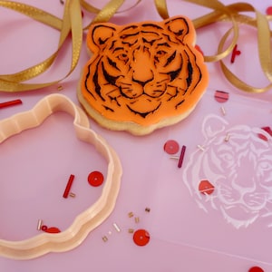 May include: An orange tiger-shaped cookie with black details, a cookie cutter, and a stencil are displayed on a pink surface. Gold ribbon and red and gold confetti add festive accents. The cookie features a detailed tiger face.