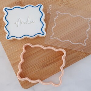 Könnte beinhalten: Ein Keks, verziert mit weißem Zuckerguss und blauem Rand, mit dem Namen "Amelia" in der Mitte. Eine transparente Kunststoffschablone und ein rosa Ausstecher mit gewelltem Rand sind ebenfalls zu sehen. Die Artikel liegen auf einem Holzschneidebrett.