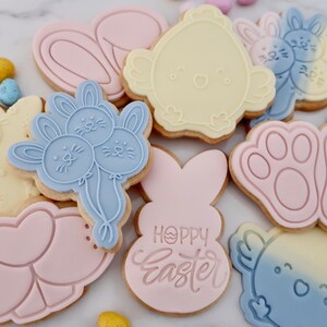 May include: Assortment of Easter-themed cookies in various shapes, including bunnies, chicks, and paw prints. The cookies are decorated with pastel pink, blue, and yellow icing. Some cookies have the words "Happy Easter" on them. Colorful candy eggs are scattered around the cookies.