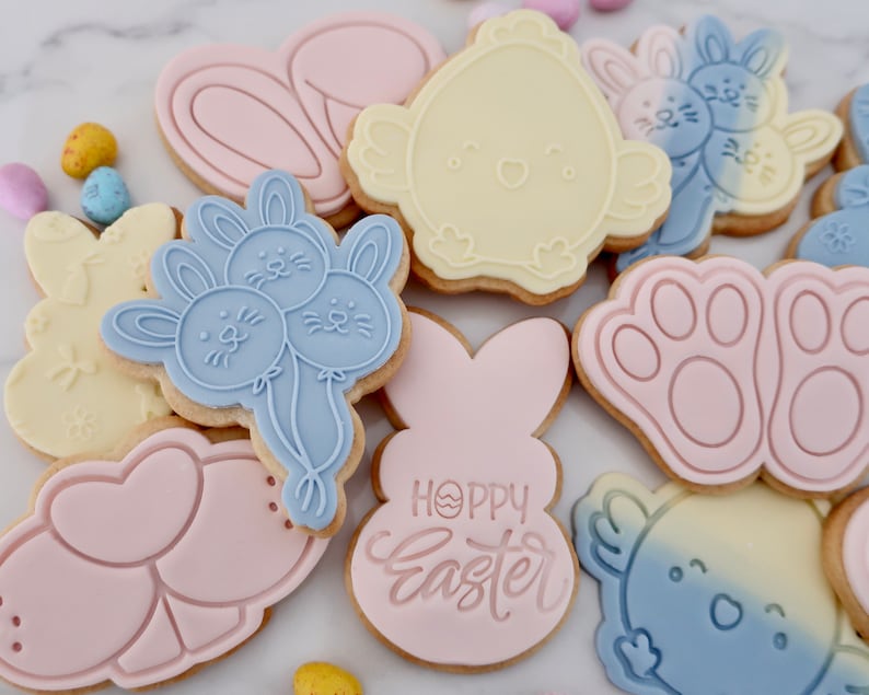 May include: Assortment of Easter-themed cookies in various shapes, including bunnies, chicks, and paw prints. The cookies are decorated with pastel pink, blue, and yellow icing. Some cookies have the words "Happy Easter" on them. Colorful candy eggs are scattered around the cookies.