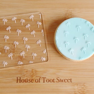 May include: A clear acrylic stamp with a square shape and a pattern of small white palm trees. A round biscuit with light blue icing and matching palm tree impressions is next to the stamp. The wooden surface has the text "House of Toot Sweet".
