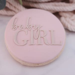 May include: A round, light pink cookie with the words "baby GIRL" in a light beige color. The cookie has a light beige edge and is set against a blurred background of beige rope and greenery.