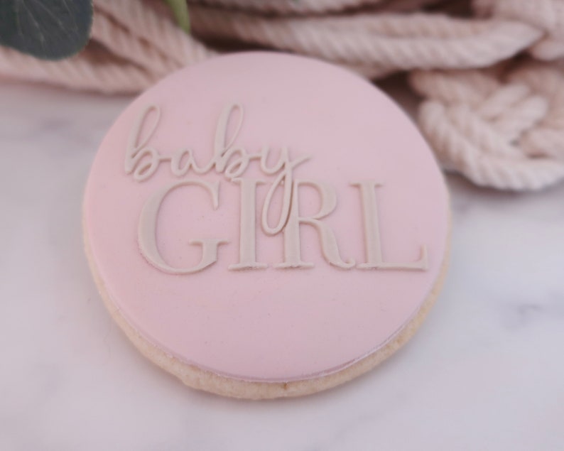 May include: A round, light pink cookie with the words "baby GIRL" in a light beige color. The cookie has a light beige edge and is set against a blurred background of beige rope and greenery.