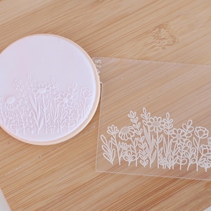 May include: A round, light pink cookie cutter with a floral design, next to a clear plastic template with the same floral pattern. The cutter is on a light brown wooden surface.