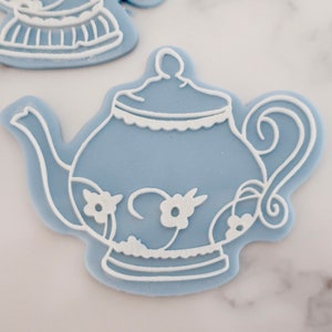 May include: Pale blue teapot-shaped cookie cutter with white floral and decorative detailing. The teapot has a handle, spout, and lid. The cookie cutter is made of plastic and is designed for baking. Other cookie cutters are visible in the background.