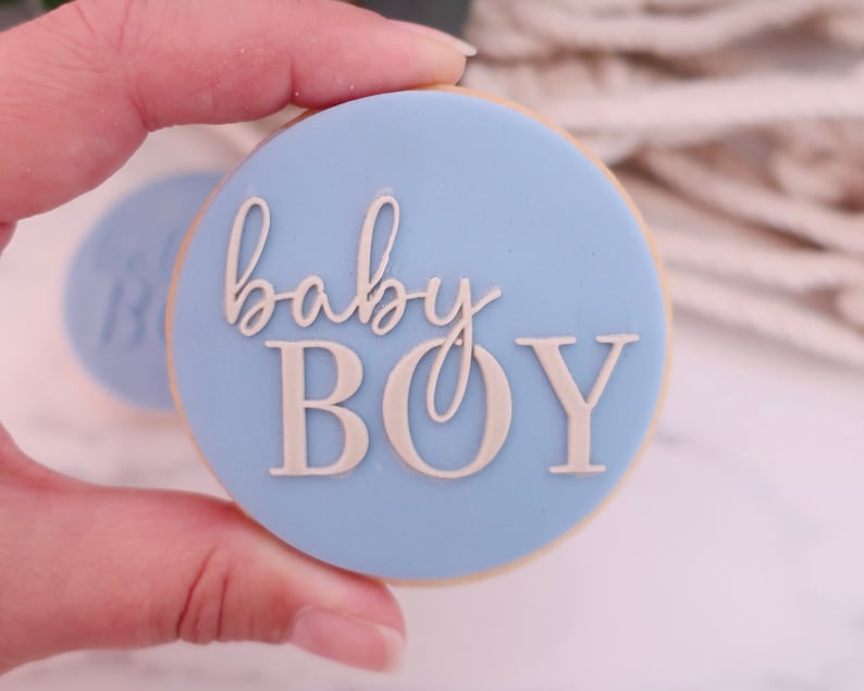 May include: A round cookie with a light blue frosting and the words "baby BOY" in a light beige color. The cookie is held in a hand, with another cookie visible in the background.