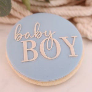 May include: A round cookie with a light blue frosting. The words "baby BOY" are written in a light beige color on the frosting. The cookie has a light beige edge.