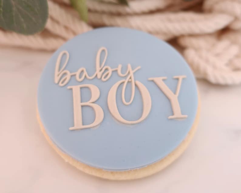 May include: A round cookie with a light blue frosting. The words "baby BOY" are written in a light beige color on the frosting. The cookie has a light beige edge.