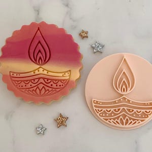 Może przedstawiać: Ozdobione ciastko i stempel do ciastek z motywem lampy diya. Ciastko ma różowo-żółty gradient ze szczegółowym wzorem lampy. Stempel jest jasnoróżowy z tym samym wzorem lampy. Rozrzuconych jest kilka posypek w kształcie gwiazdek.