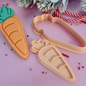 May include: A carrot-shaped biscuit with a green top, alongside two carrot-shaped biscuit cutters. The biscuit has three curved lines and sits on a light base. The cutters are a pale peach colour. The background is a light purple.