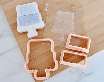 May include: A set of light peach plastic cookie cutters and a clear acrylic embosser plate featuring a two-tiered cake design. The embosser plate displays a cake silhouette with the text HOTS and includes a light blue cake-shaped cookie piece.